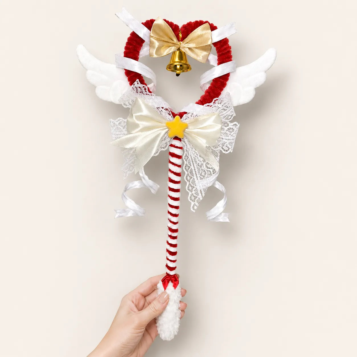 A hand holds a decorative wand with a red-and-white striped handle, white angel wings, a red heart-shaped top, a gold bow, lace details, and a small bell.