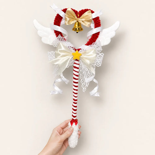 A hand holds a decorative wand with a red-and-white striped handle, white angel wings, a red heart-shaped top, a gold bow, lace details, and a small bell.