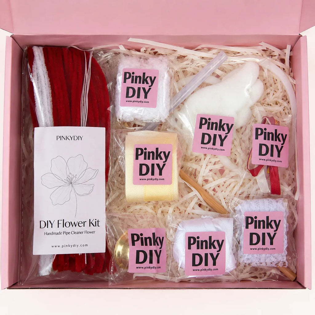 A Pinky DIY DIY Flower Kit gift box containing a red pipe cleaner bundle, a white bunny-shaped item, a yellow soap bar, and several packaged flower-shaped items on a pink box interior with shredded paper.