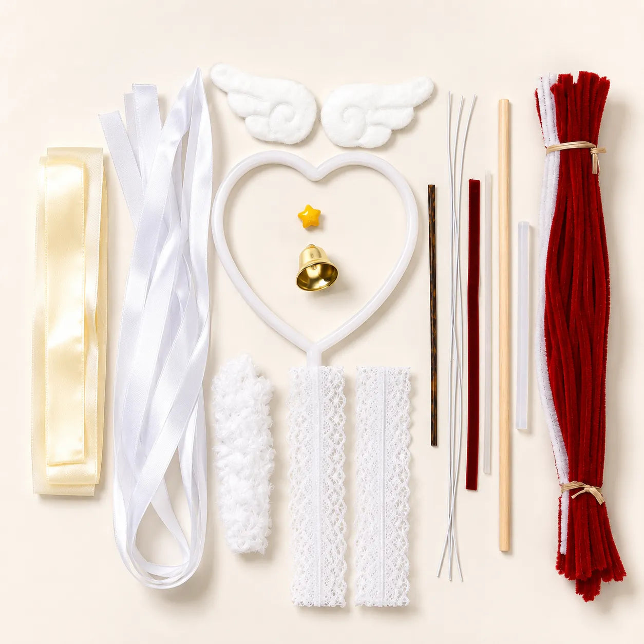 Angel-themed craft kit laid out on a light beige background, including white angel wings, a white heart-shaped wand, white lace strips, a white fluffy piece, a cream ribbon, red and white pipe cleaners, wooden sticks, and a small gold bell.