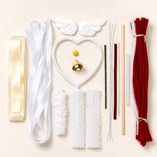Angel-themed craft kit laid out on a light beige background, including white angel wings, a white heart-shaped wand, white lace strips, a white fluffy piece, a cream ribbon, red and white pipe cleaners, wooden sticks, and a small gold bell.