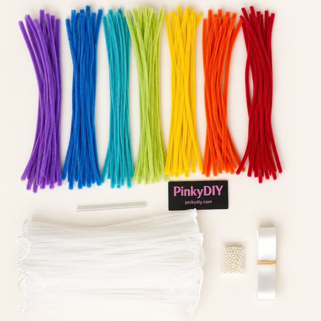 A DIY craft kit laid out on a white background, featuring six vertical rows of colorful pipe cleaners in purple, blue, teal, green, yellow, orange, and red. Below the pipe cleaners are white tulle sheets, a small white bead bundle, a white ribbon, and a black brand label that reads 'PinkyDIY pinkydiy.